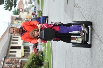 Ein Mann und ein junges Mädchen fahren auf einem schwarzen Segway auf einer asphaltierten Straße, mit Gebäuden und Grünflächen im Hintergrund.