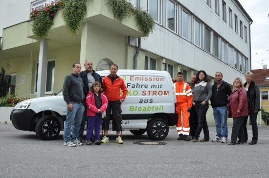 Eine Gruppe von Menschen steht vor einem weißen Van mit grünem Text, der 'Fahr mit 0 Emission aus Bioabfall' lautet. Sie alle lächeln und scheinen für ein Foto zu posieren.