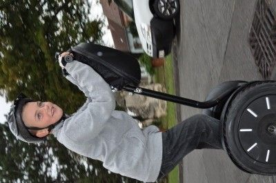 Ein junger Junge fährt einen schwarzen Segway auf einer asphaltierten Straße, trägt einen Helm und ein graues Sweatshirt. Der Segway hat einen schwarzen Lenker und ein großes weißes Rad.