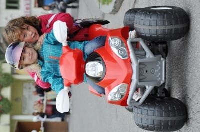 Zwei junge Mädchen fahren an einem sonnigen Tag auf einem roten Spielzeug-ATV, eine trägt einen Helm und die andere ein rotes Jackett.