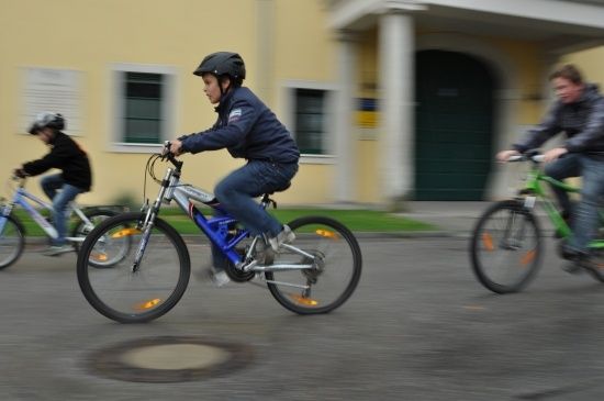 Drei Radfahrer fahren vor einem gelben Gebäude eine Straße hinunter, einer trägt einen schwarzen Helm.