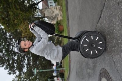 Eine junge Person fährt einen schwarzen Segway auf einer asphaltierten Straße, trägt einen Helm und ein graues Sweatshirt. Bäume und ein Gebäude sind im Hintergrund zu sehen.