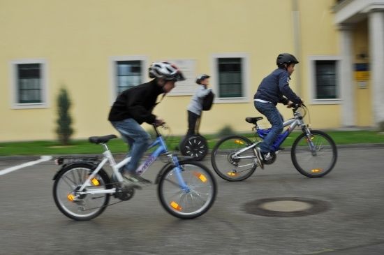 Zwei Jungen fahren auf einem Parkplatz mit Fahrrädern, einer trägt einen Helm, der andere einen Rucksack. Ein dritter Junge fährt im Hintergrund auf einem Einrad.
