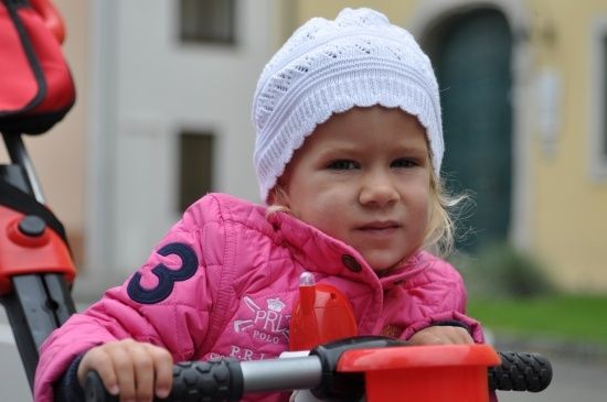 Ein kleines Mädchen in einer rosa Jacke und einer weißen Mütze fährt an einem sonnigen Tag mit einem roten Roller.