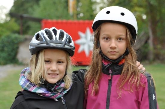 Zwei junge Mädchen mit Helmen und Jacken stehen zusammen in einem Park mit rotem Hintergrund.
