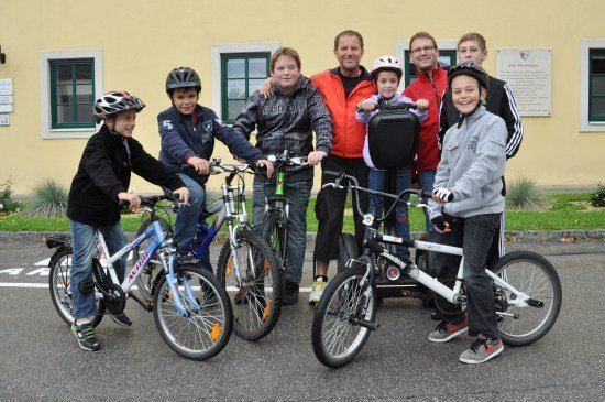 Eine Gruppe von Menschen, darunter Kinder und Erwachsene, steht vor einem Gebäude mit gelben Wänden und grünen Fenstern und posiert für ein Foto mit ihren Fahrrädern.