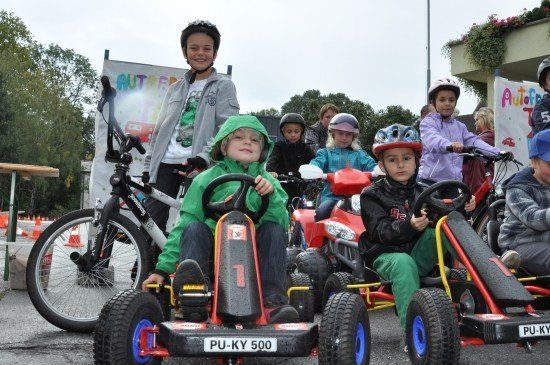 Eine Gruppe von Kindern, die Helme tragen, fährt in einer Reihe auf einem asphaltierten Weg mit Pedal-Go-Karts. Im Hintergrund befindet sich ein Banner mit bunter Beschriftung.