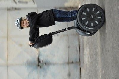 Ein junger Mensch mit Helm fährt einen schwarzen Segway auf einer Betonfläche. Der Segway hat ein großes rundes Rad und die Person hält die Lenkstange.