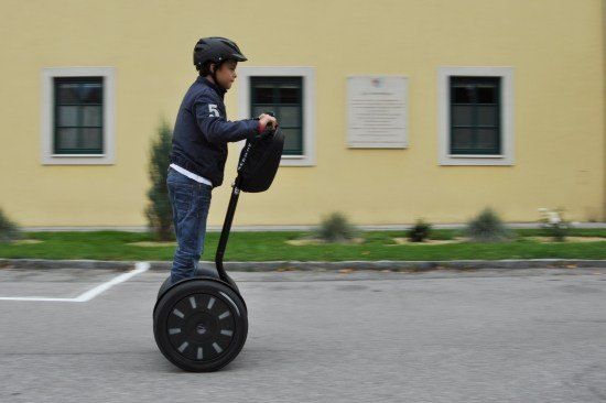 Ein junger Junge fährt einen Segway auf einer asphaltierten Straße vor einem gelben Gebäude mit weißen Fensterrahmen.