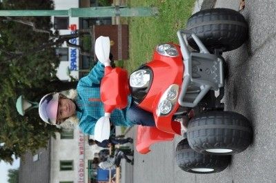 Ein junges Mädchen fährt auf einer asphaltierten Straße mit einem roten vierrädrigen motorisierten Spielzeug und trägt einen Helm und eine blaue Jacke. Bäume und ein Gebäude mit einem Schild sind im Hintergrund.