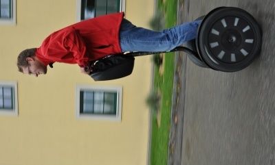 Ein Mann in einem roten Jackett und blauen Jeans fährt einen Segway auf einer Straße mit einem gelben Gebäude im Hintergrund.