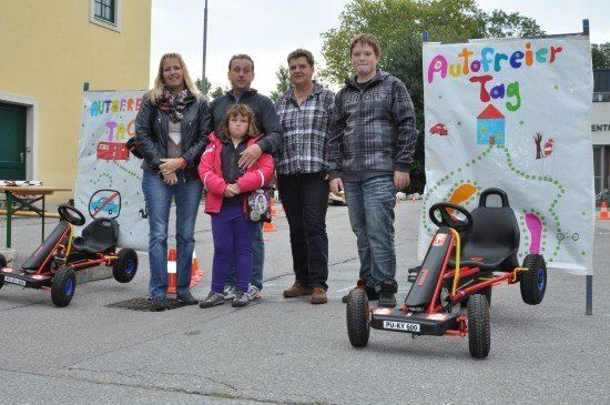 Eine Familie mit fünf Personen steht vor einem Gebäude mit zwei Go-Karts, eines davon ist mit 'Autofreier Tag' beschriftet. Die Familienmitglieder lächeln und scheinen für ein Foto zu posieren.