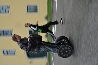 Ein Mann fährt einen Segway, während eine weitere Person hinter ihm steht, beide tragen schwarze und grüne Jacken. Sie befinden sich auf einer asphaltierten Straße vor einem gelben Gebäude mit mehreren Fenstern.