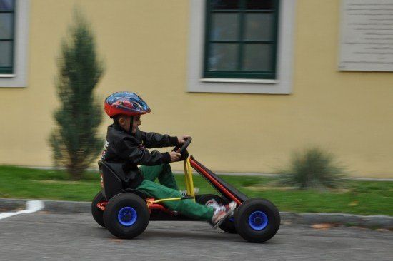 Ein junger Junge mit Helm fährt einen Go-Kart die Straße hinunter, mit einem gelben Gebäude im Hintergrund.