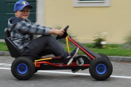 Ein junger Junge mit einer blauen Kappe fährt einen roten Go-Kart mit blauen Rädern auf einer Straße.