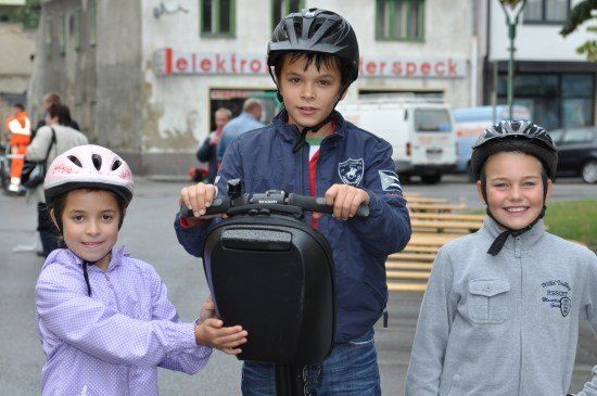 Drei Kinder stehen auf einer Straße und tragen Helme. Der Junge in der Mitte hält einen schwarzen Segway. Hinter ihnen befinden sich Bänke und ein Gebäude mit einem Schild, das 'Elektro Speck' besagt.