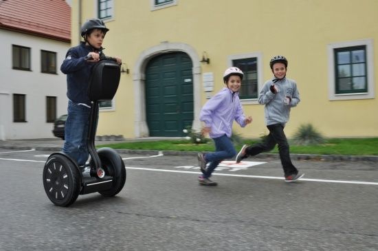 Drei Kinder sind draußen, eines fährt einen Segway, während die anderen nebenher laufen. Sie stehen vor einem gelben Gebäude mit grünen Türen und Fenstern.