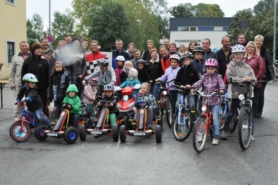 Eine Gruppe von Kindern und Erwachsenen posiert für ein Foto mit ihren Fahrrädern und Go-Karts in einer Außenumgebung.