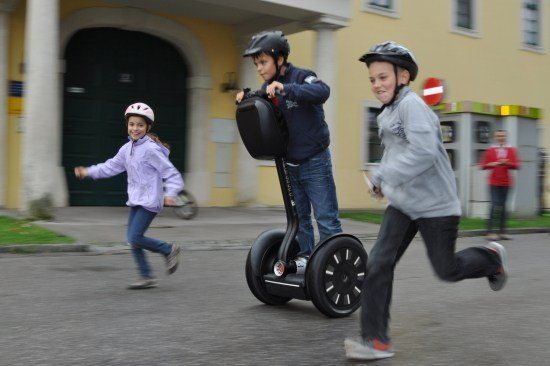 Drei Kinder fahren auf einer Straße mit einem Segway, wobei der Junge in der Mitte einen Helm trägt und das Lenkrad hält. Das Mädchen links und der Junge rechts laufen neben dem Segway. Im Hintergrund befindet sich ein Gebäude mit einer Garagentür und einem Stoppschild.
