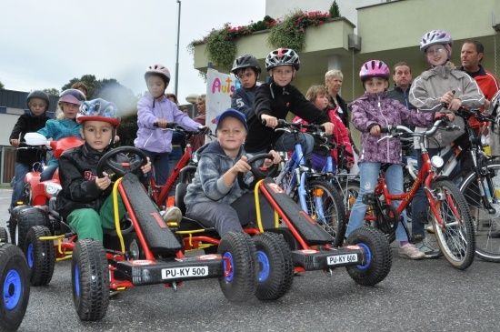 Eine Gruppe von Kindern mit Helmen fährt auf Pedal-Go-Karts in einem Parkplatz. Einige Erwachsene stehen in der Nähe.