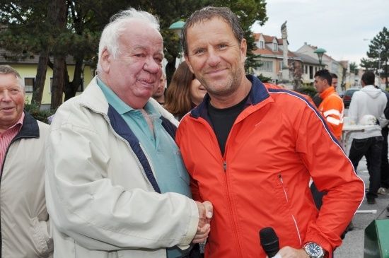 Zwei Männer, einer in einer weißen Jacke und der andere in einer orangefarbenen Jacke, schütteln sich die Hände vor einer Menge. Der Mann in der orangefarbenen Jacke hält ein Mikrofon.