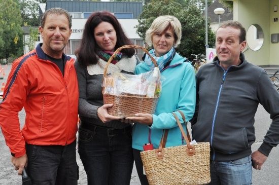Vier Personen stehen vor einem Gebäude, zwei Frauen halten einen Korb und eine Handtasche, ein Mann im roten Jackett lächelt, und ein anderer im grauen Jackett.