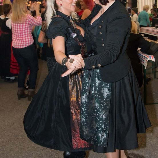 Zwei Frauen tanzen bei einem Festival zusammen, eine trägt ein schwarzes Kleid und die andere eine schwarze Jacke über einem Blumenkleid. Hinter ihnen tanzen und genießen die Leute das Event.