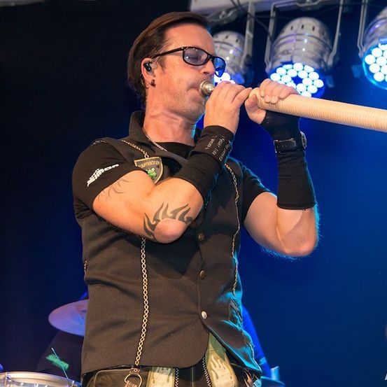 Ein Mann mit Tätowierungen an den Armen spielt auf der Bühne eine lange Trompete mit einer Band hinter ihm.