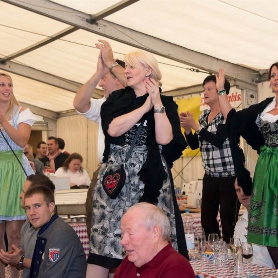 Eine Gruppe von Menschen in traditioneller deutscher Kleidung klatscht und jubelt in einem Zelt, wobei eine Frau in der Mitte ein schwarzes Kleid und eine herzförmige Tasche trägt.