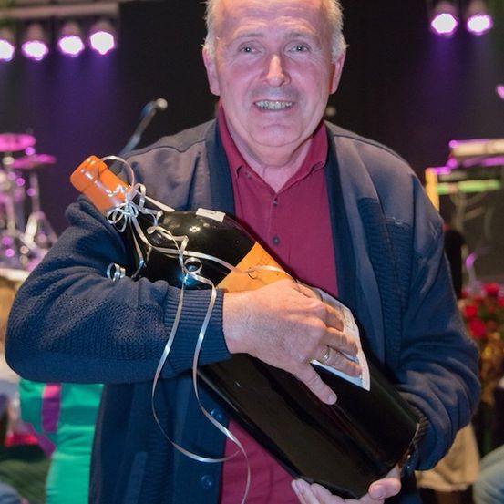 Ein älterer Mann hält eine große Flasche Wein, lächelt und trägt ein rotes Hemd und eine blaue Jacke. Der Hintergrund zeigt eine Bühne mit Musikinstrumenten und lila Lichtern.