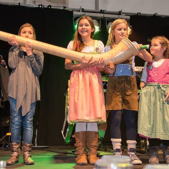 Vier Mädchen in traditioneller Kleidung stehen auf der Bühne, halten ein großes Horn und lächeln für die Kamera.