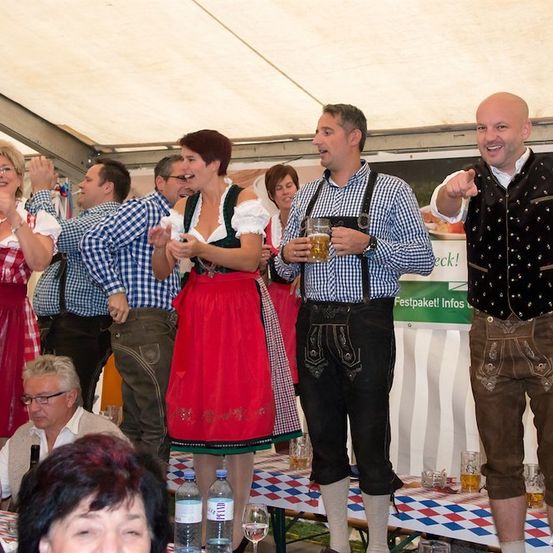 Eine Gruppe von Menschen in traditioneller bayerischer Kleidung versammelt sich unter einem Zelt, lächelt und hält Getränke in den Händen. Ein Mann in einem blau karierten Hemd und einer suspender hält ein Glas Bier. Ein anderer Mann in einer schwarzen Weste zeigt auf etwas. Ein Tisch mit Flaschen und Gläsern steht im Vordergrund.