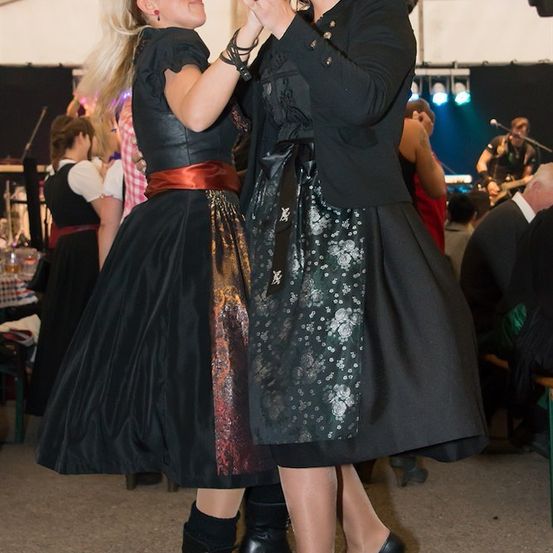 Zwei Frauen in traditioneller Kleidung tanzen bei einer lebhaften Veranstaltung zusammen, mit einem Gitarristen und anderen Gästen im Hintergrund.
