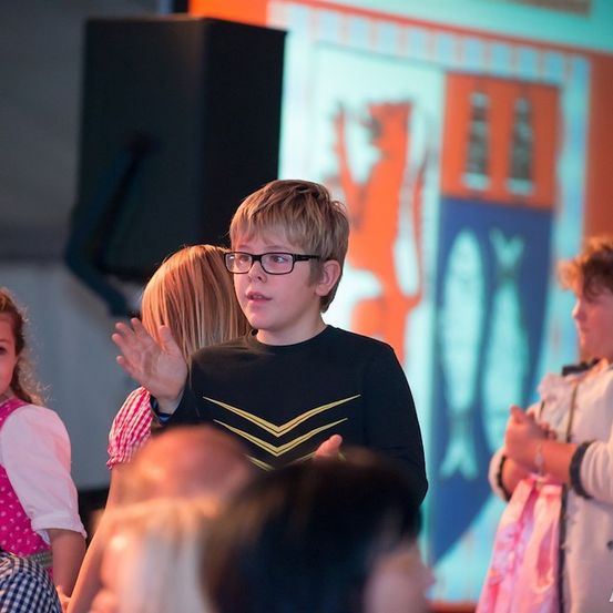 Eine Gruppe von Kindern und Erwachsenen ist drinnen versammelt, wahrscheinlich bei einer Veranstaltung. Ein Junge mit Brille klatscht und schaut nach oben. Hinter ihm zeigt eine große Leinwand ein Bild einer Katze.
