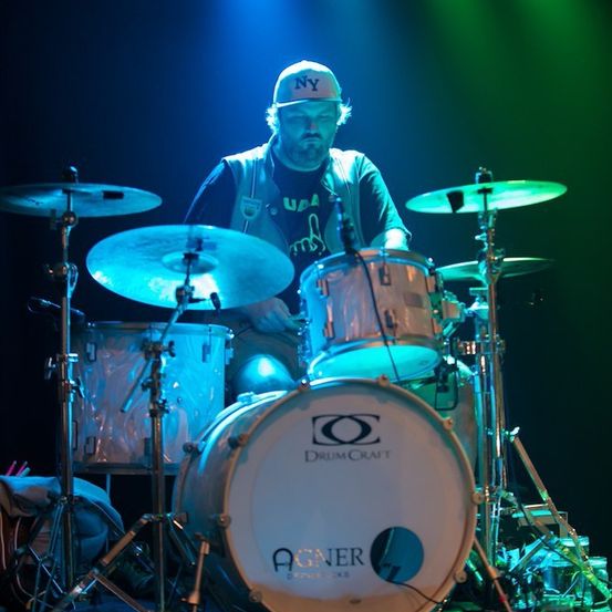 Ein Schlagzeuger mit einer New Yorker Kappe spielt an einem Schlagzeug mit einem DrumCraft-Bassdrum in einem dunklen Raum mit blauen und grünen Lichtern.