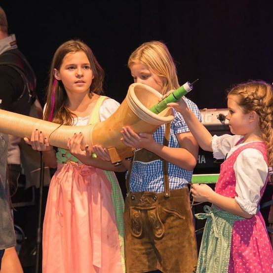 Vier Kinder in traditioneller bayerischer Kleidung halten ein langes Horninstrument auf einer Bühne.