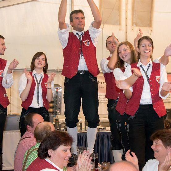 Eine Gruppe von Menschen in traditioneller Kleidung führt auf der Bühne auf, einer der Männer steht auf einem Stuhl. Das Publikum klatscht und lächelt.