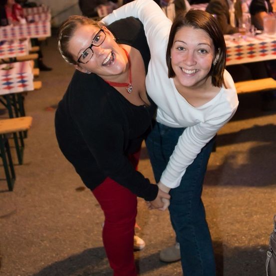 Zwei Frauen lächeln und posieren für ein Foto bei einer Outdoor-Veranstaltung, eine trägt eine Brille und eine Halskette, die andere ein weißes Shirt. Hinter ihnen sind Tische mit rot, weiß und blau karierten Tischdecken aufgestellt.