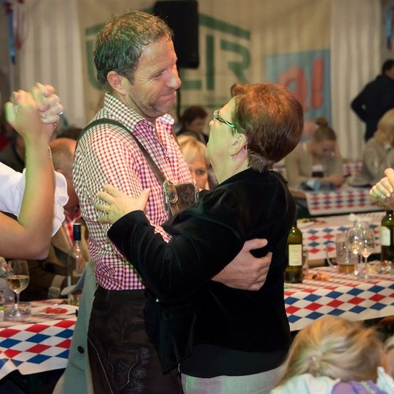 Ein Mann und eine Frau tanzen bei einer Party zusammen, umgeben von anderen Leuten, die an Tischen mit Weinflaschen und Gläsern sitzen.