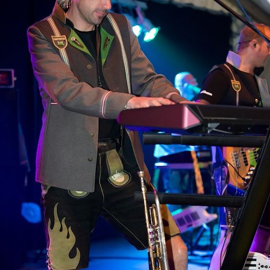 Ein Mann in traditioneller bayerischer Kleidung spielt auf der Bühne ein Keyboard, vor ihm steht eine Trompete. Ein anderer Mann spielt Gitarre im Hintergrund.