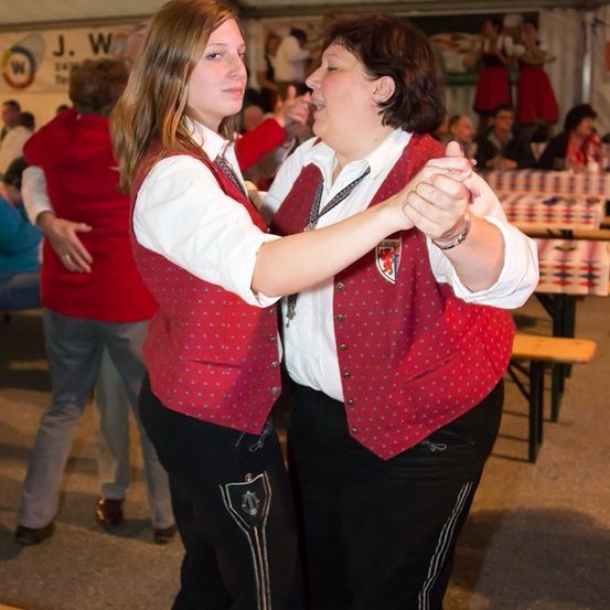 Zwei Frauen in roten Westen und schwarzen Hosen tanzen zusammen bei einer Veranstaltung im Freien, mit anderen Leuten und Tischen im Hintergrund.