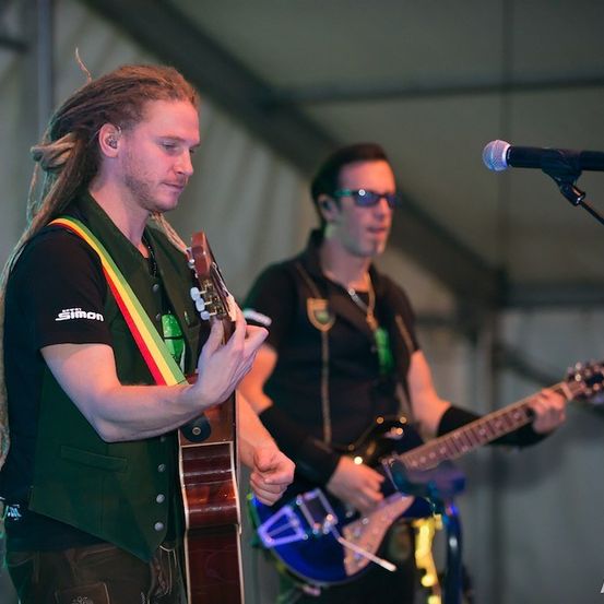 Zwei Männer spielen auf der Bühne mit Gitarren. Ein Mann hat Dreadlocks und spielt eine braune Gitarre, während der andere Sonnenbrille trägt und eine blaue Gitarre spielt. Ein Mikrofon steht vor ihnen.