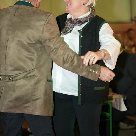 Ein älteres Paar tanzt bei einer Veranstaltung zusammen, die Frau trägt einen Schal und der Mann einen braunen Mantel.
