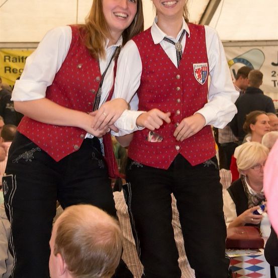 Zwei Frauen in traditioneller Kleidung, die lächelnd für ein Foto bei einer Veranstaltung im Freien posieren. Sie tragen rote Westen mit weißen Kragen und Krawatten. Hinter ihnen sitzen und stehen Leute, mit einem Banner im Hintergrund.