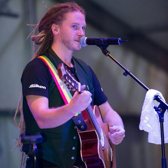 Ein Mann mit Dreadlocks, der eine grüne Weste und ein schwarzes Shirt mit einem Regenbogenband trägt, spielt Gitarre und singt in ein Mikrofon auf der Bühne.
