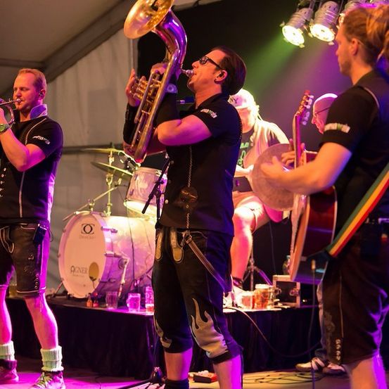 Eine Band spielt auf der Bühne mit einem Trompeter, einem Tubaspieler, einem Schlagzeuger und einem Gitarristen. Die Bühne ist mit lila und grünen Lichtern beleuchtet.