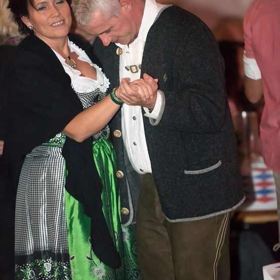 Ein Mann und eine Frau tanzen bei einem Festival zusammen, beide in traditioneller Kleidung. Die Frau trägt ein grünes Kleid mit einem weißen und schwarzen Karomuster. Der Mann trägt einen grauen Mantel mit goldenen Knöpfen und ein weißes Hemd.