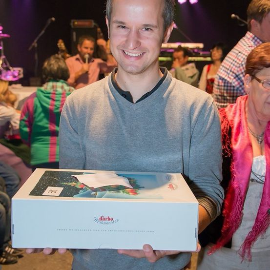 Ein Mann hält eine Box mit festlichem Design, lächelt in die Kamera, mit Menschen im Hintergrund.