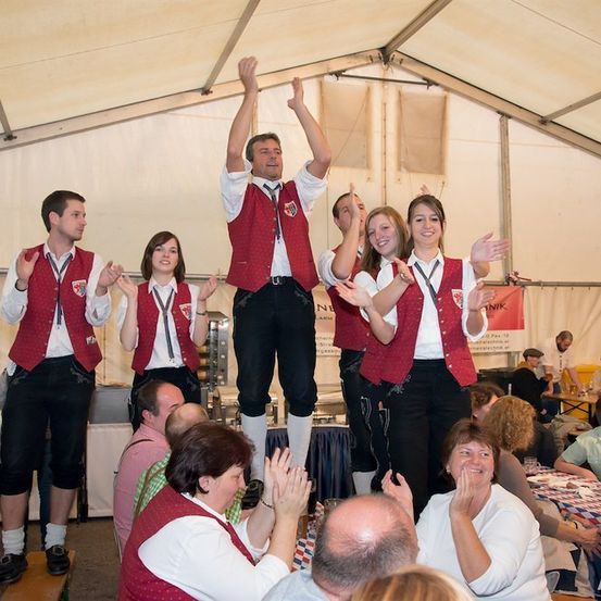 Eine Gruppe von Menschen in traditioneller Kleidung tritt auf einer Bühne unter einem Zelt auf, während ein jubelndes Publikum im Hintergrund zusieht.