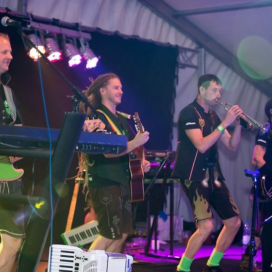Eine Band spielt auf einer Bühne mit verschiedenen Musikinstrumenten, darunter Trompete, Gitarre und Keyboard. Die Musiker sind in Freizeitkleidung gekleidet und scheinen ihre Aufführung zu genießen.